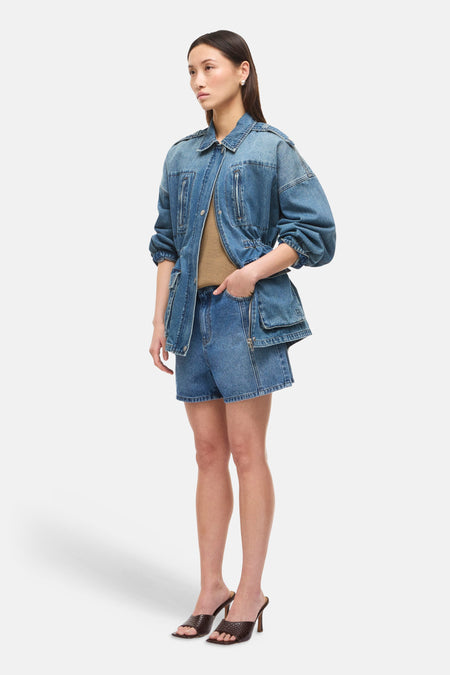 Veste courte en jean bleu moyen de ROUGE EDIT, avec fermeture éclair, poches zippées, cordon de serrage, portée avec un short en denim et des mules marron.