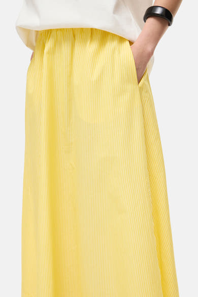 Jupe longue jaune de ROUGE EDIT à fines rayures verticales blanches, portée avec un T-shirt blanc et un bracelet noir, main dans la poche.