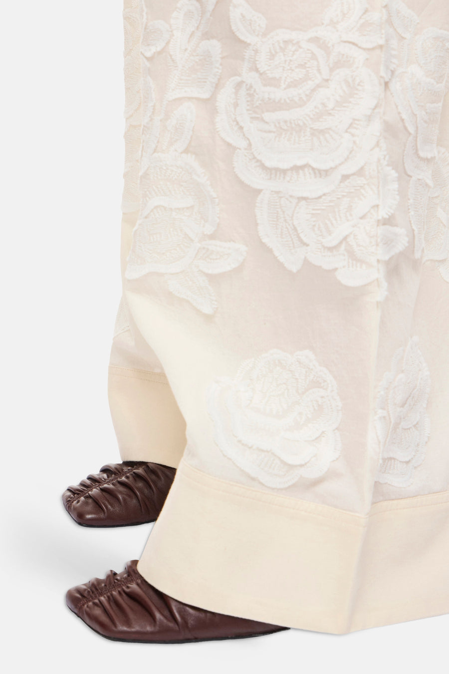 Pantalon beige de ROUGE EDIT à broderies florales blanches en relief et larges revers, porté avec des chaussures plates marron.