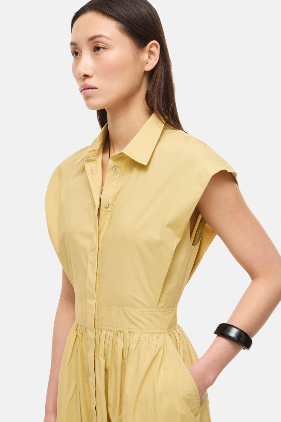 Robe chemise jaune de ROUGE EDIT, à col, sans manches, taille froncée, poches latérales, portée de profil avec un bracelet noir.