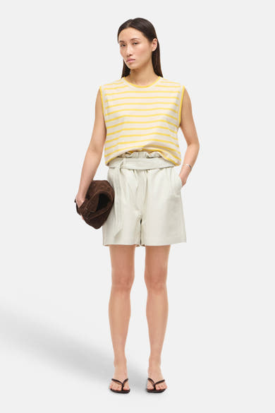 Top sans manches rayé jaune et écru de ROUGE EDIT, porté avec un short gris clair et des tongs marron, tenant un sac en daim.