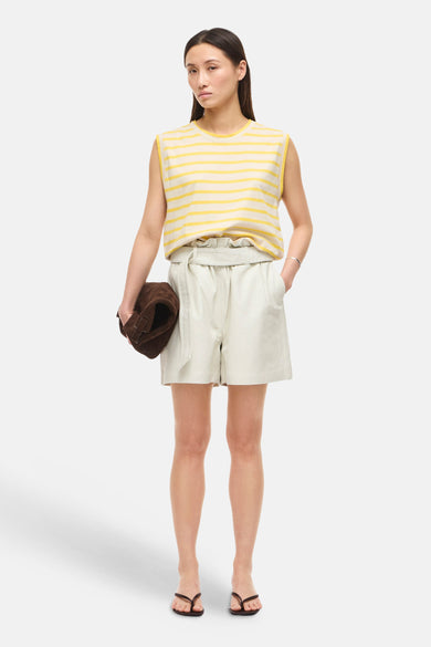 Top sans manches rayé jaune et écru de ROUGE EDIT, porté avec un short gris clair et des tongs marron, tenant un sac en daim.