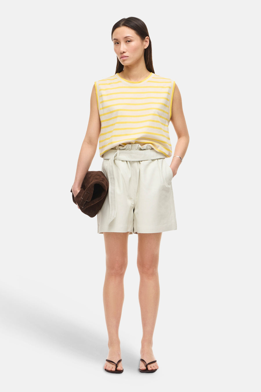 Top sans manches rayé jaune et écru de ROUGE EDIT, porté avec un short gris clair et des tongs marron, tenant un sac en daim.