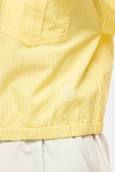 Chemisier jaune à manches courtes de ROUGE EDIT, à rayures blanches fines, avec une poche poitrine, un logo brodé et un ourlet élastiqué, porté sur un pantalon blanc.