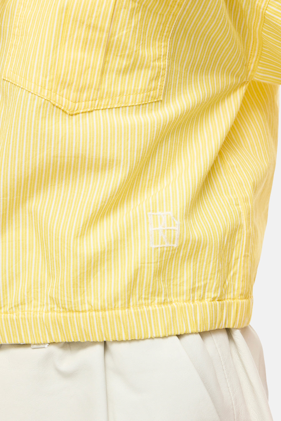Chemisier jaune à manches courtes de ROUGE EDIT, à rayures blanches fines, avec une poche poitrine, un logo brodé et un ourlet élastiqué, porté sur un pantalon blanc.