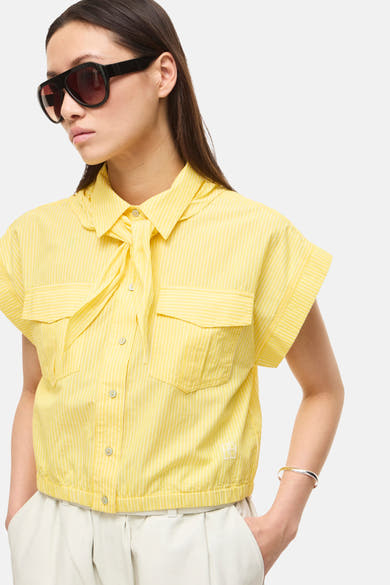 Chemisier à manches courtes jaune de ROUGE EDIT avec fines rayures blanches, col noué et poches poitrine, porté avec un pantalon écru.