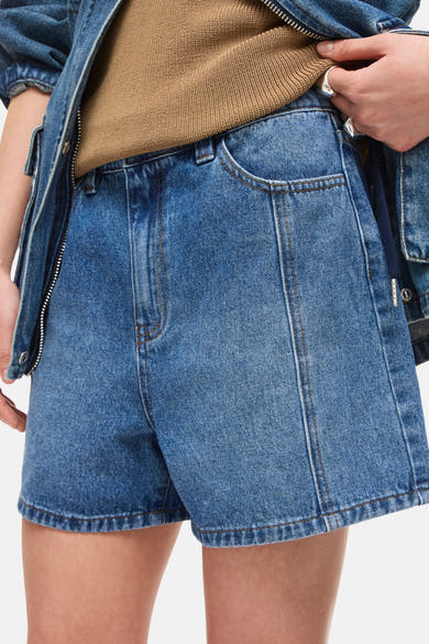 Short en jean bleu moyen de ROUGE EDIT, avec poche et coutures apparentes, porté avec un pull tricoté beige et une veste en jean.