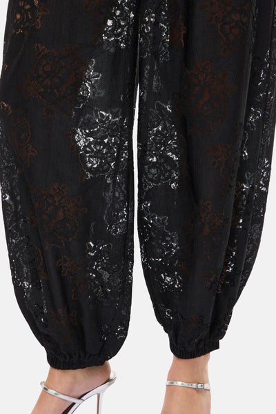 Pantalon noir en dentelle par Vila, avec motif floral décoratif et chevilles élastiquées, porté avec des sandales argentées.