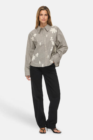 Beige checkered long-sleeve blouse with white floral embroidery, worn by model with black wide-leg pants and black sandals, front view.