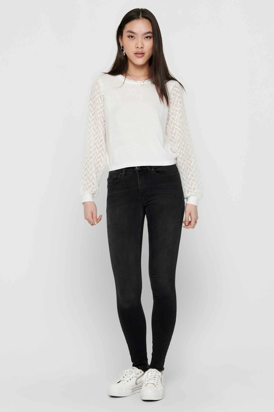 Zwarte skinny jeans van ONLY, gedragen met een witte top met lange doorschijnende mouwen en witte schoenen.