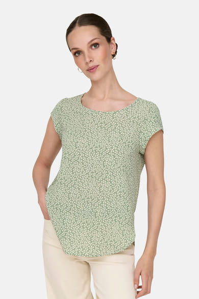 Chemisier vert à manches courtes par ONLY, imprimé de motifs végétaux, porté avec un pantalon clair et des boucles d'oreilles dorées.