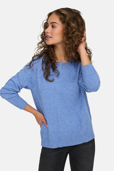 Pull à col rond bleu de ONLY®, porté avec un jean noir et un collier argenté, modèle avec les cheveux bouclés.