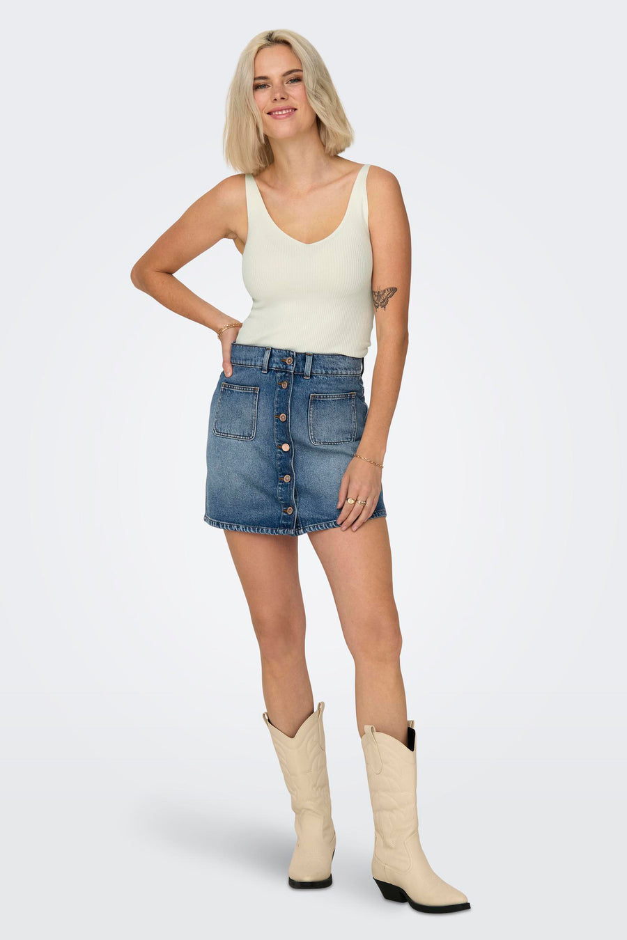 Débardeur blanc de JDY, porté avec une jupe en jean boutonnée et des bottes de cow-boy.