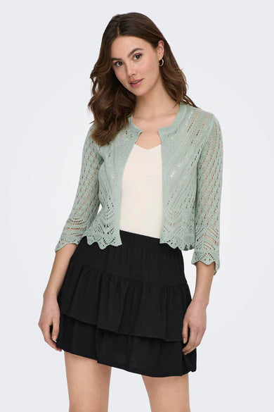 Gilet vert à maille ajourée de JDY, porté sur un débardeur blanc et une jupe noire à volants.

