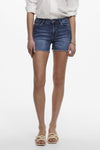 Short bleu en jean de ONLY, avec ourlet effiloché, porté avec une chemise blanche et des sandales.