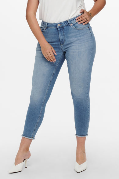 Jean skinny bleu moyen de CARMAKOMA, coupe cheville avec ourlet effiloché, porté avec des escarpins blancs.
