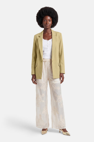 Blazer vert de GOA GOA, porté ouvert sur un top blanc et un pantalon brodé crème avec des talons dorés.