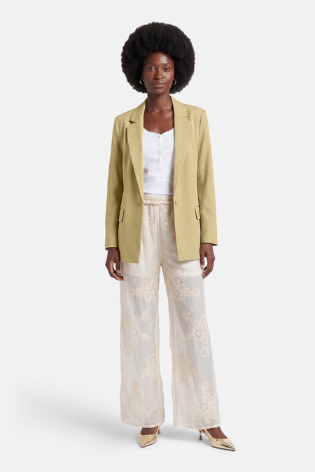 Blazer vert de GOA GOA, porté ouvert sur un top blanc et un pantalon brodé crème avec des talons dorés.