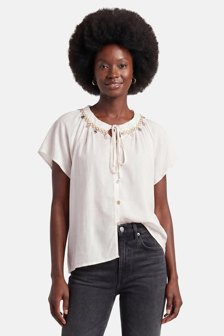 Witte GOA GOA blouse, ronde hals versierd met een gouden ketting en kleurrijke kwastjes, korte wijde mouwen, gedragen met een donkere jeans.