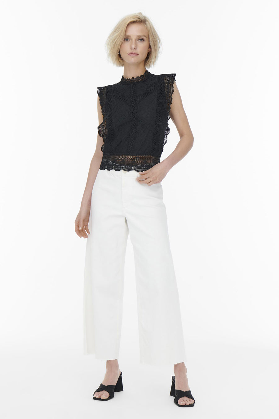 Blouse noire sans manches de ONLY, en dentelle délicate, portée avec un pantalon blanc et des mules à talons.

