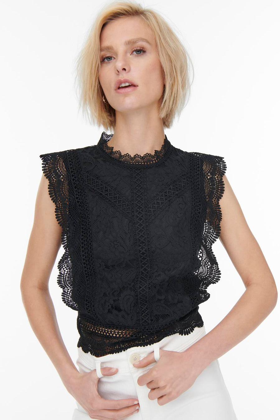 Blouse noire en dentelle de ONLY®, à col montant et à bordures festonnées, portée avec un pantalon blanc.
