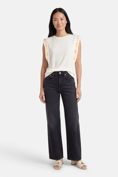 Beige T-shirt van GOA GOA met ronde hals, met geborduurde ruches op de schouders, gedragen met een grijze jeans en beige muiltjes.