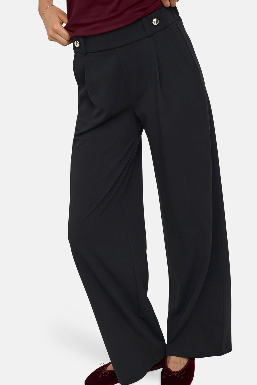 Black wide-leg elastic waist trousers with front pleats and silver buttons, side view on model wearing burgundy top and velvet shoes