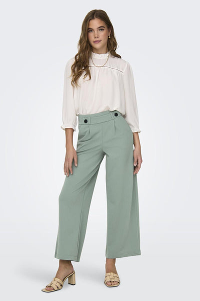 Pantalon costume vert par JDY, avec taille haute et coupe large, porté avec un chemisier blanc et des sandales.
