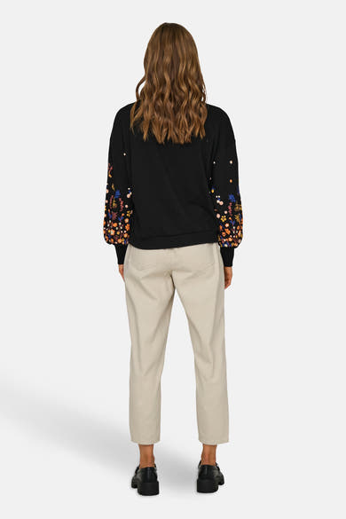 Sweat noir de ONLY®, avec des manches longues fleuries, porté avec un pantalon beige et des chaussures noires.