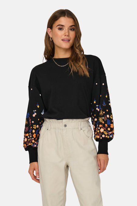 Zwarte trui van ONLY, met lange ballonmouwen geborduurd met kleurrijke bloemenpatronen; gedragen met een beige broek met hoge taille en een fijne gouden ketting.