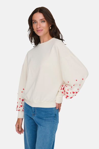 Ecru sweatshirt van ONLY®, met lange mouwen versierd met kleine rode hartjes, gedragen met een blauwe jeans.