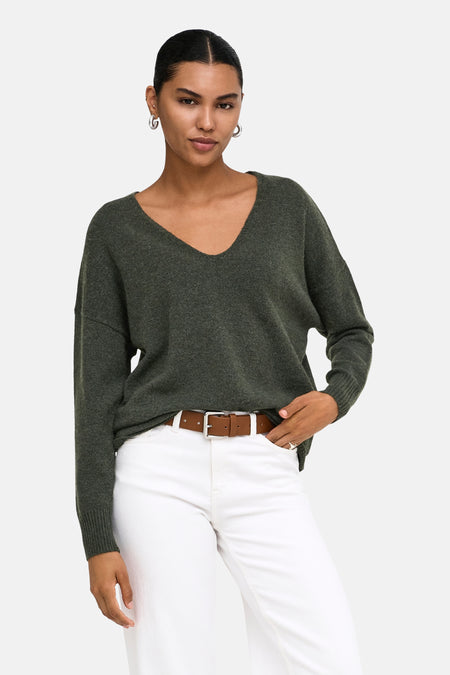 Pull col V vert foncé en maille douce par ONLY®, porté avec un pantalon blanc et une ceinture marron, vue de face.