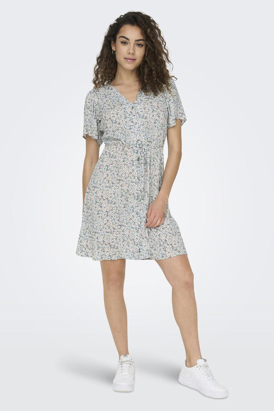 Groene jurk met korte mouwen van ONLY® met bloemenprint, V-hals en strikdetail in de taille, gedragen met witte sneakers.