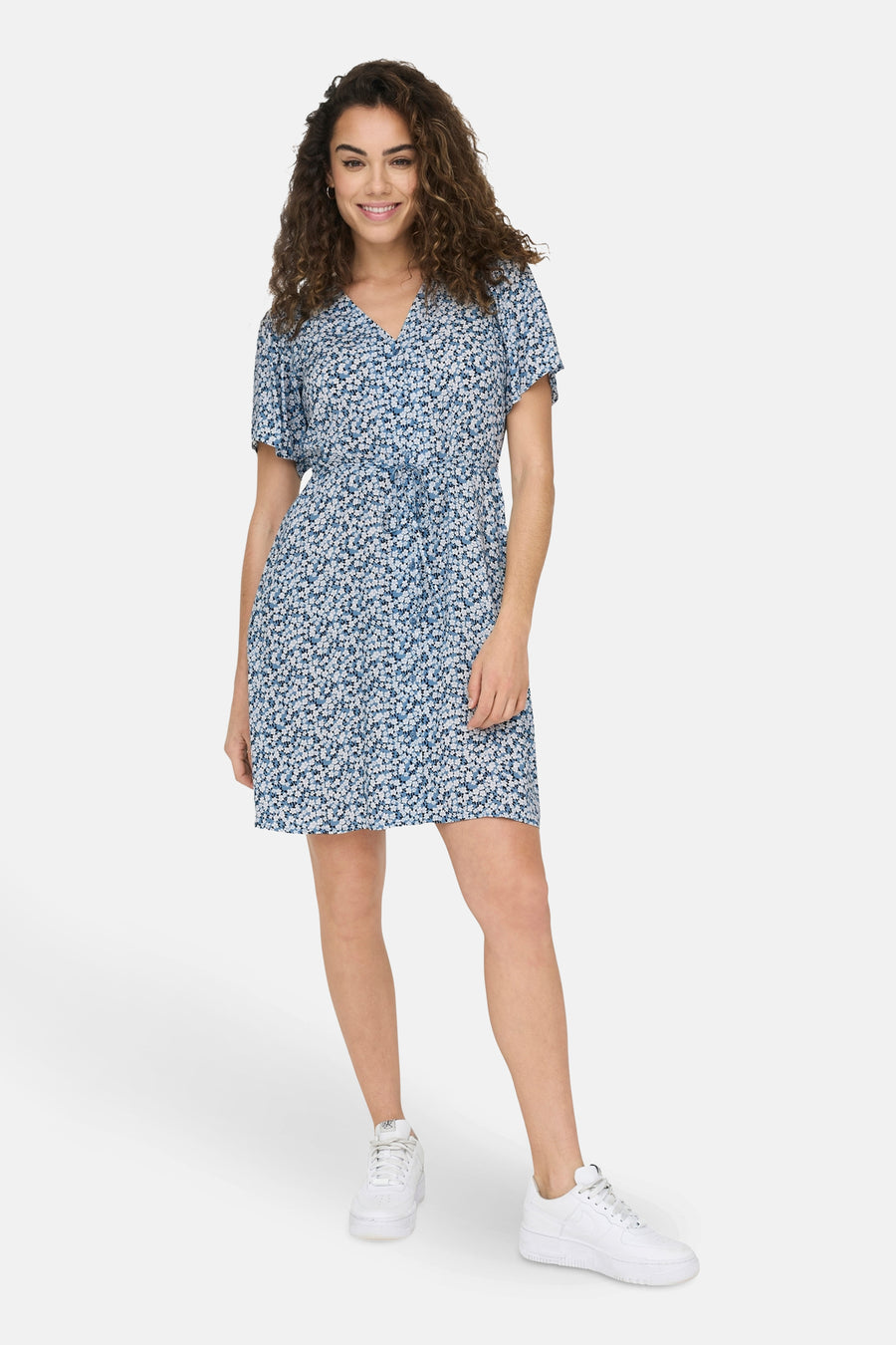Blauwe jurk met korte mouwen van ONLY® met bloemenprint en strikceintuur, gedragen met witte sneakers.