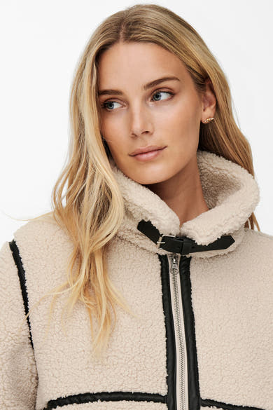 Veste longue écru de JDY avec garniture noire et col en fausse fourrure, portée avec des boucles d'oreilles dorées.