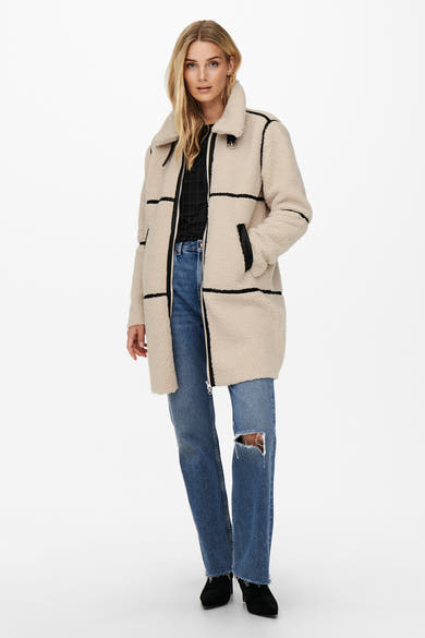 Longue veste écru de JDY, avec un col et des bordures contrastées noires, portée sur un jean bleu déchiré.
