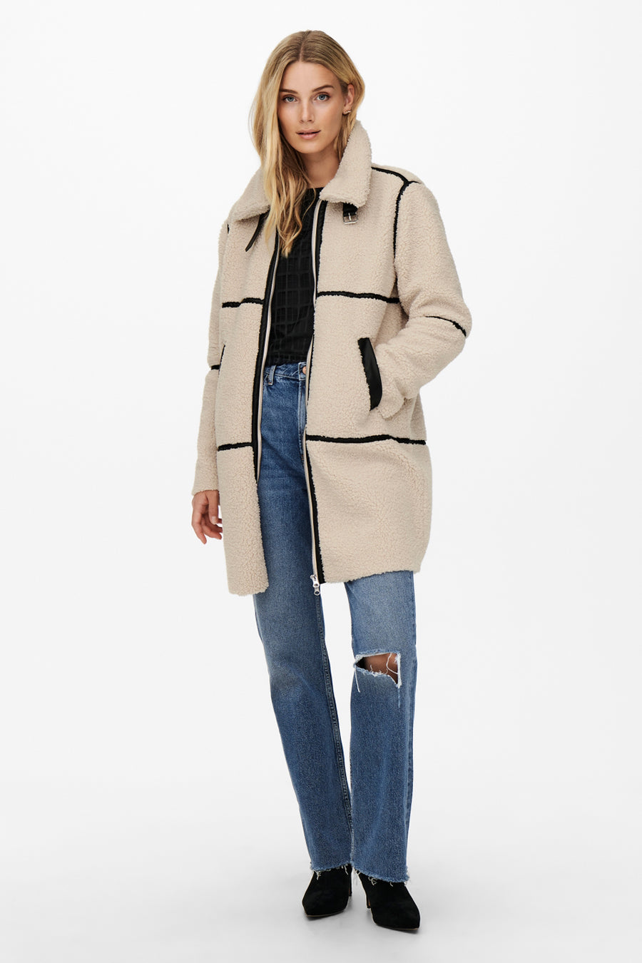 Longue veste écru de JDY, avec un col et des bordures contrastées noires, portée sur un jean bleu déchiré.
