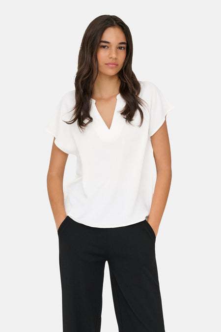 Blouse blanche de JDY à col en V, manches courtes amples, au tissu légèrement texturé, portée avec un pantalon noir.