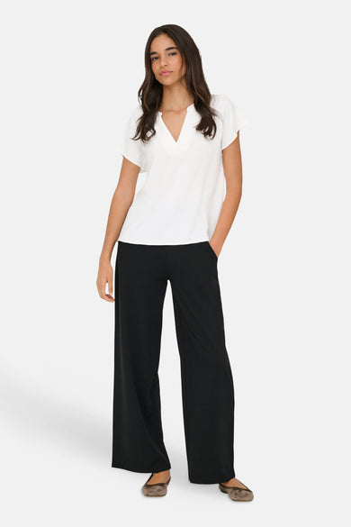 Blouse blanche de JDY à manches courtes et col en V, portée avec un pantalon noir ample et des ballerines marron.