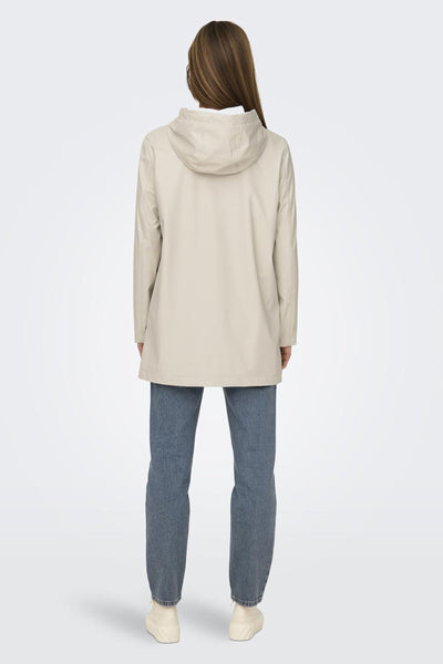 Ecru regenjas van ONLY, met een capuchon, gedragen met een blauwe jeans en beige sneakers.