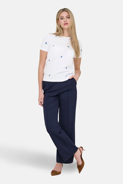 T-shirt à manches courtes blanc de ONLY®, orné de motifs floraux bleus, porté avec un pantalon bleu marine et des escarpins marron.
