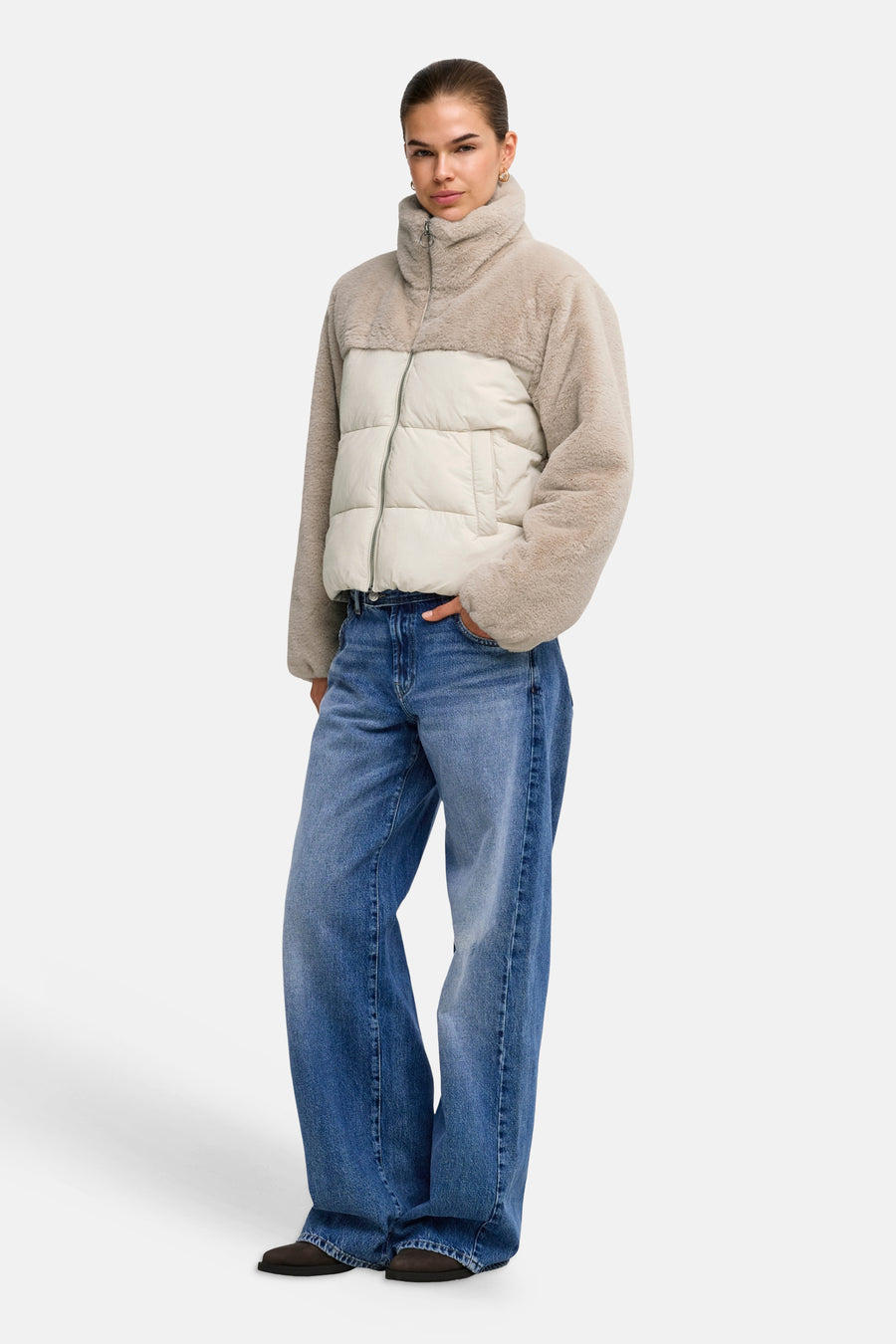 Beige and cream two-tone quilted puffer jacket with plush turtleneck and long sleeves, worn with blue wide-leg jeans and dark shoes, front view.
