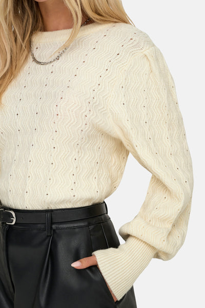Pull beige à col rond de ONLY®, avec motif ajouré et manches bouffantes, porté avec un pantalon noir.
