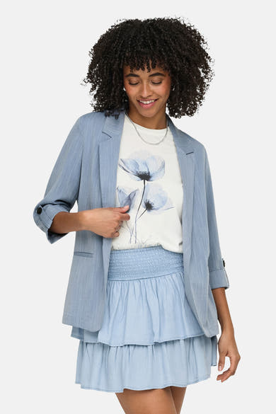 Blauwe ONLY® blazer, driekwartmouwen opgerold, gedragen over een T-shirt met bloemenprint en een bijpassende rok.