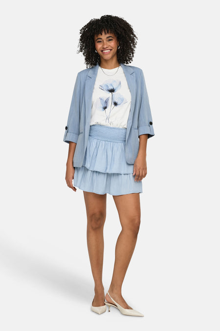 Blazer bleu ONLY®, manches trois-quarts retroussées, porté sur un t-shirt à motif floral et une jupe assortie.