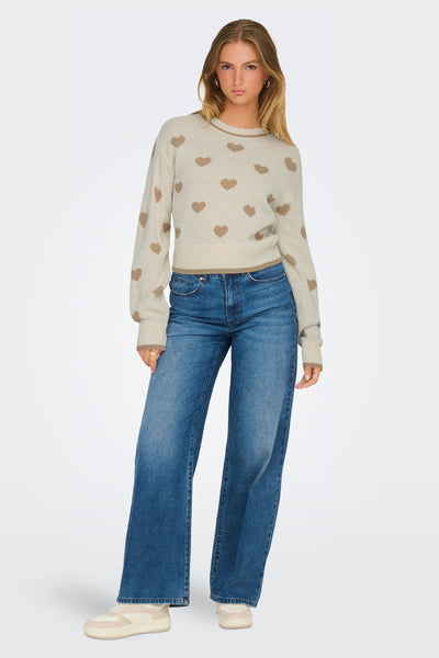 Pull à col rond beige de JDY, à motif de coeurs marron, porté avec un jean bleu large et des baskets beiges.
