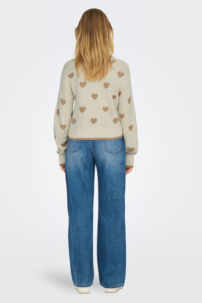 Pull à col rond beige de JDY, orné de motifs de cœurs marron, porté avec un jean bleu.
