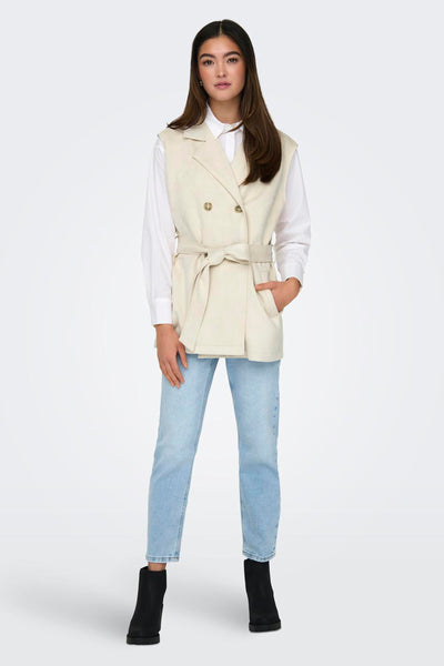 Veste beige sans manches de ONLY, portée sur une chemise blanche et un jean bleu clair, avec des bottines noires.