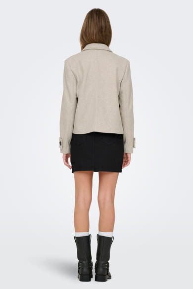 Korte beige jas van ONLY, van achteren gezien, gedragen met een zwarte rok en zwarte laarzen.