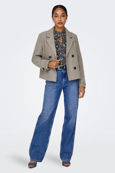 Veste courte beige de ONLY, avec double boutonnage et portée avec un chemisier à motifs et un jean.
